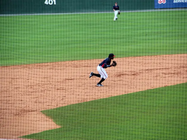 The image shows a baseball player wearing a cap and gloves, running to first base during a game. In...