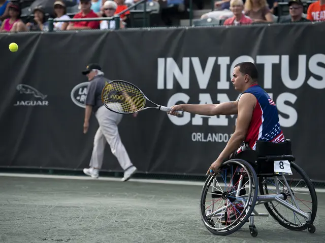 The image shows a man in a wheelchair playing tennis on a court. He is holding a tennis racket in...