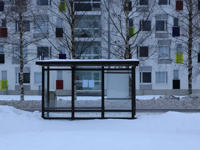 The image shows a bus stop in the middle of a snowy street, surrounded by trees and a building with...