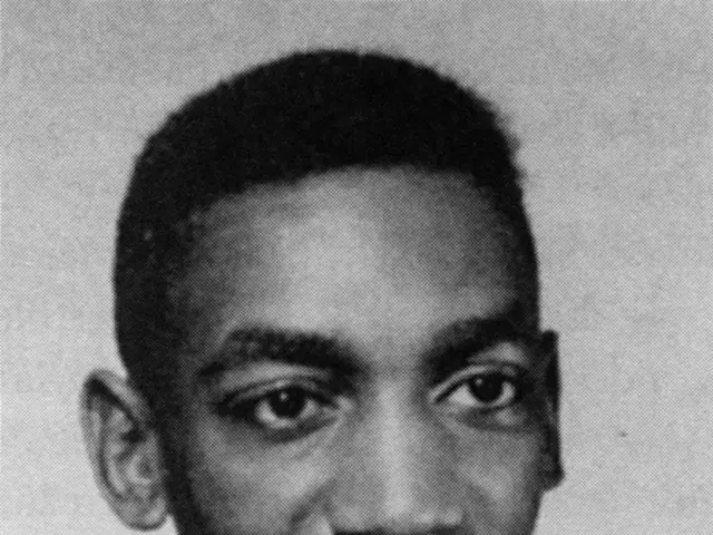 The image shows a black and white photo of a man in a basketball uniform, smiling. He is wearing a...