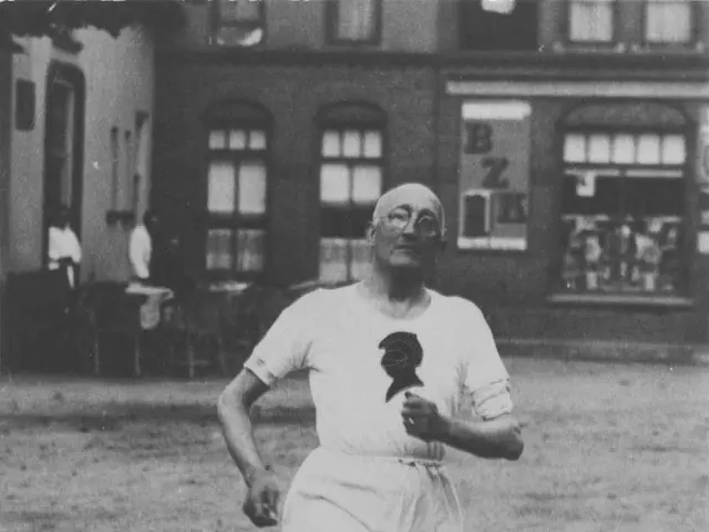 The image shows an old black and white photo of a man running in a race, with a building in the...