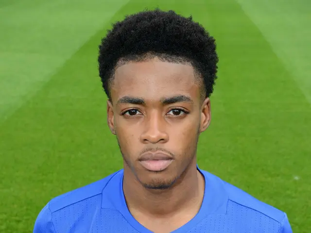 The image shows a young man in a blue t-shirt standing on top of a soccer field. He is wearing a...
