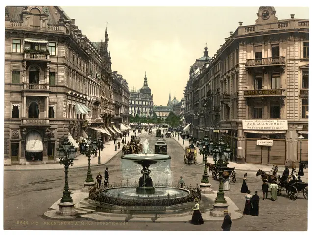 The image shows an old photo of a city street with a fountain in the center. There are many people...