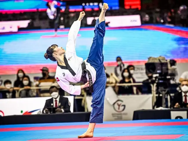 The image shows a woman in a karate kimono doing a kick in front of a crowd of people, some of whom...