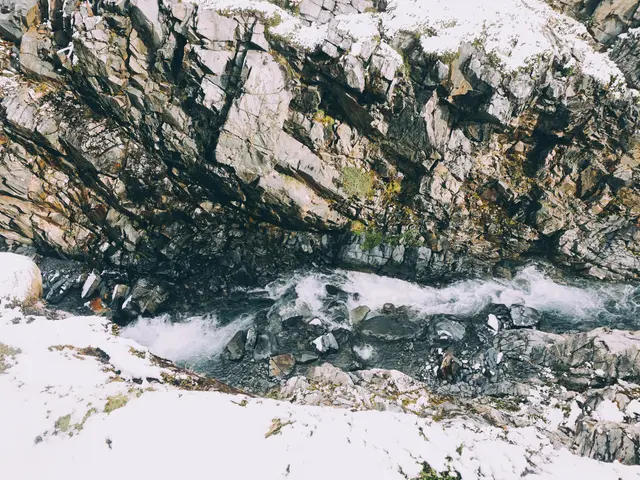 The image shows a small stream running through a rocky mountain side covered in snow. The water is...