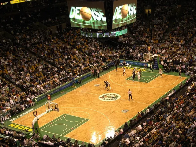 The image shows a basketball game being played in a large arena, with a group of people sitting in...