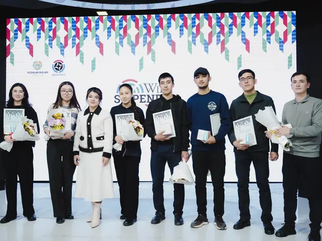 The image shows a group of people standing next to each other on a stage, holding books and flower...