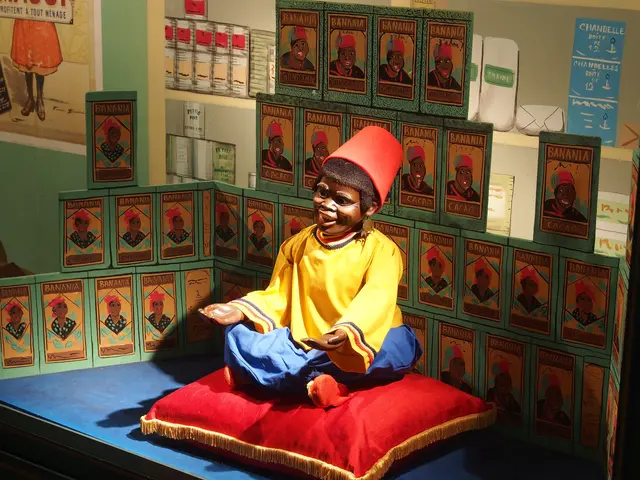 The image shows a young boy sitting on top of a red pillow on a stage, wearing a yellow and blue...
