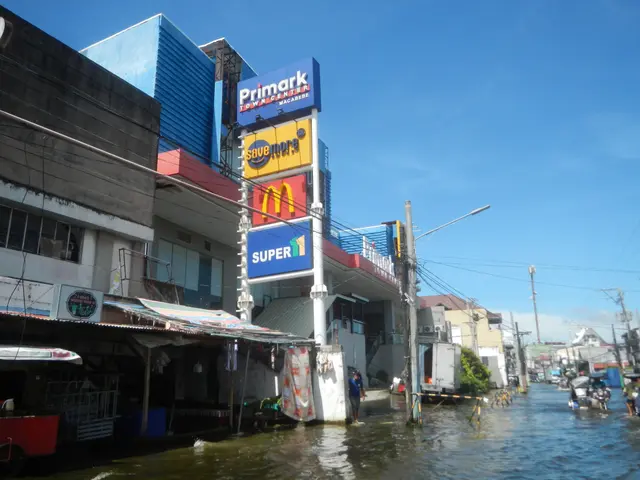 The image shows a flooded street in the middle of a city, with buildings, hoardings, electric...