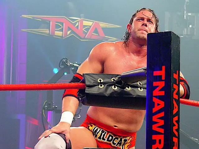 The image shows a man standing in a wrestling ring with his hands on his knees, surrounded by ropes...