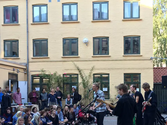 The image shows a group of people sitting on the ground in front of a building, with some of them...