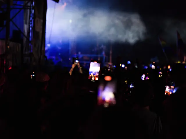 The image shows a crowd of people at a concert with their cell phones in hand, illuminated by the...