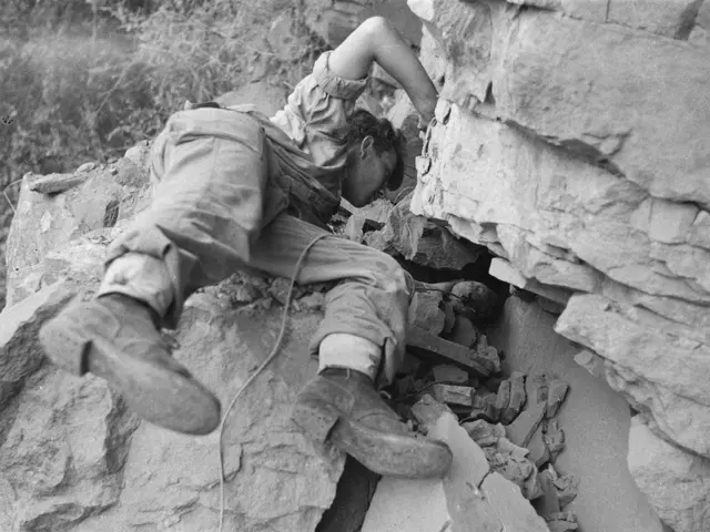 The image shows a man climbing up the side of a cliff, surrounded by trees in the background. He is...