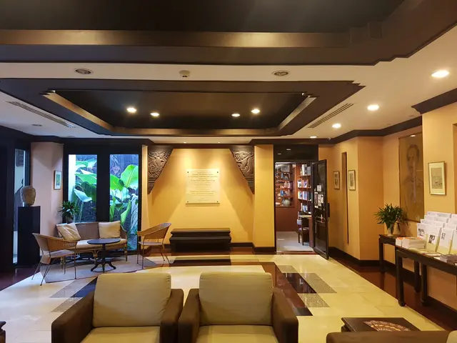 The image shows the lobby of a hotel with couches, chairs, tables, houseplants, books arranged in...