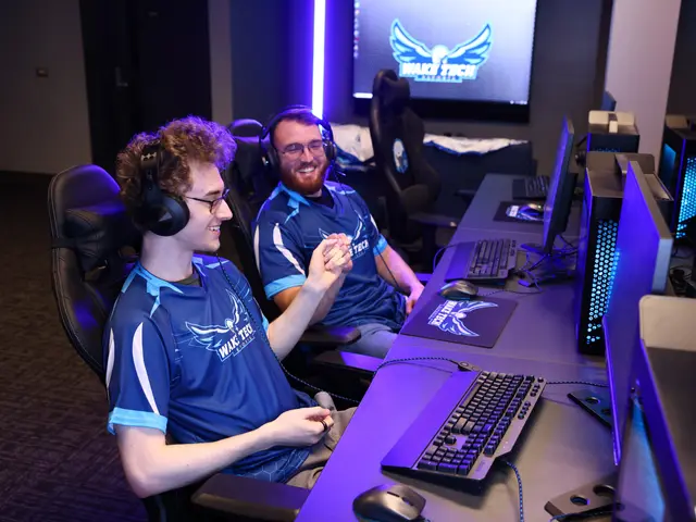 The image shows two men sitting at a desk in front of a computer, wearing blue t-shirts and...