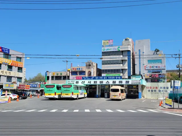 The image shows a city street with buses parked on the side of it, surrounded by buildings with...