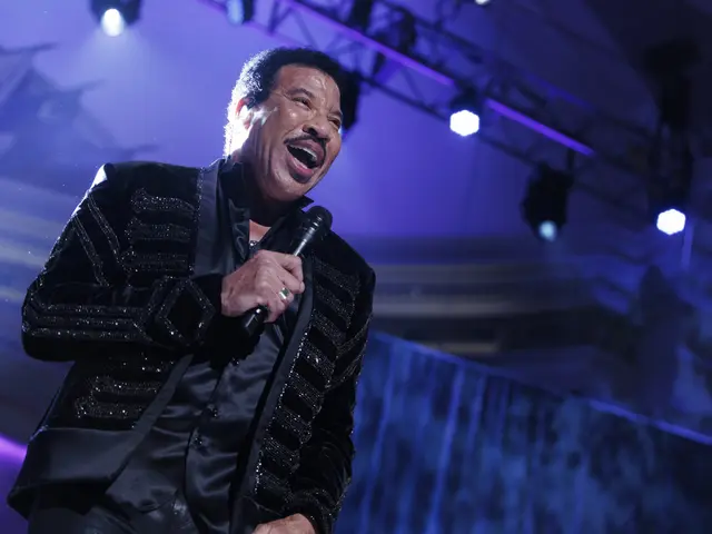 The image shows Lionel Richie performing at the Grammys, wearing a black dress and holding a...