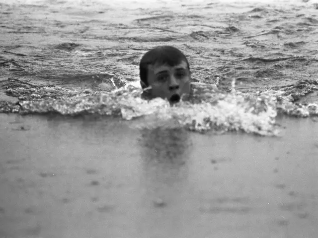 The image shows a young boy swimming in the ocean with his mouth open, surrounded by sand at the...