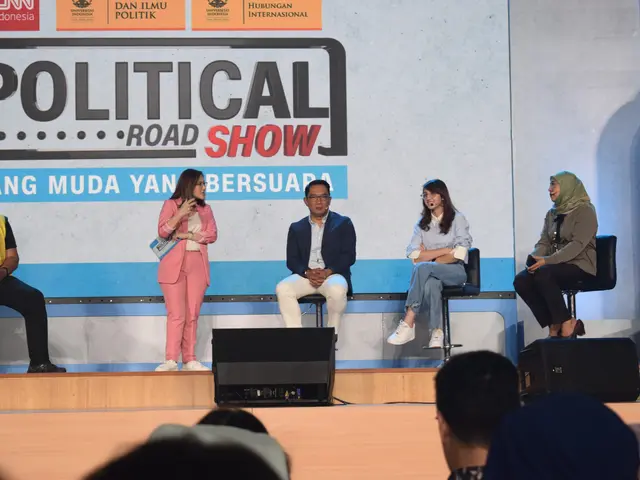 The image shows a group of people sitting on top of a stage, with a woman holding a microphone in...