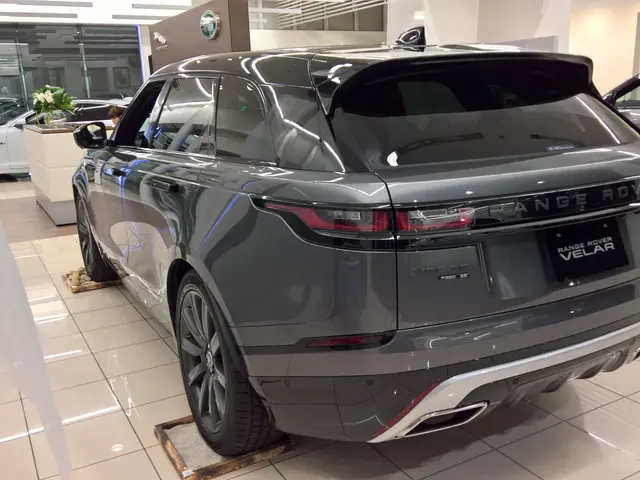 The image shows a grey Range Rover Velar parked in a showroom, surrounded by other cars on the...