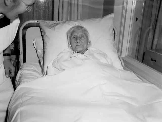 The image shows an elderly woman laying in a hospital bed with a nurse standing beside her. The bed...