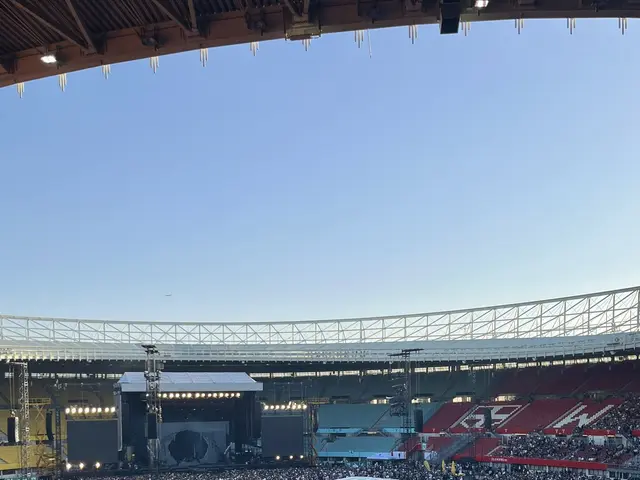 The image shows a large crowd of people sitting in a stadium, with some standing, surrounded by...