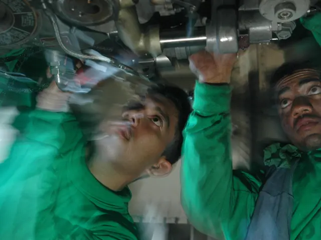 The image shows two men in green shirts working on a car engine in a factory. They are holding...