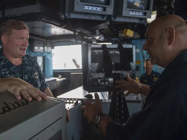 The image shows two men in military uniforms talking to each other in front of a computer,...