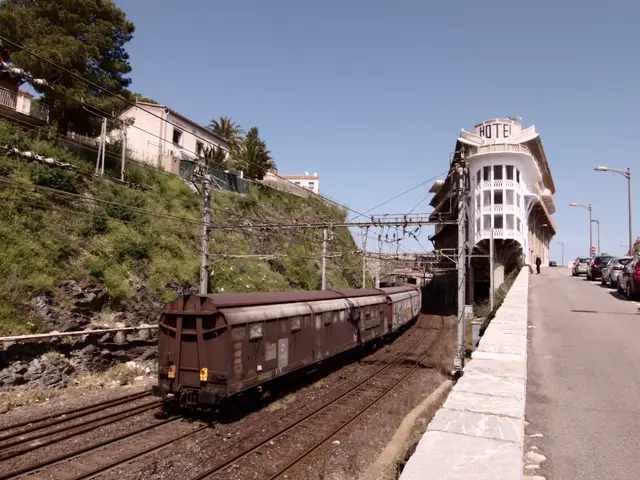 The image shows a freight train traveling down train tracks next to a building, with a platform on...