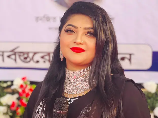 The image shows a woman in a black sari holding a microphone, with a banner and flowers in the...