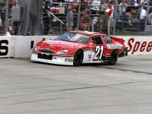 The image shows a NASCAR car driving down a race track in front of a crowd of people, some of whom...