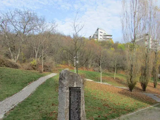 The image shows a monument in the middle of a park with a plaque on it, surrounded by grass, dried...
