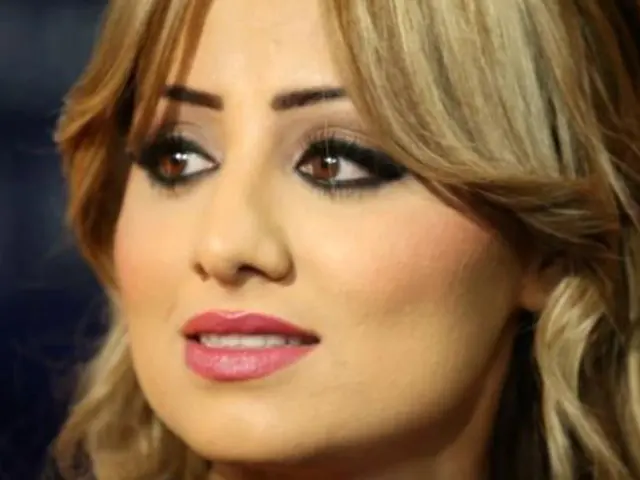 The image shows a woman with long blonde hair and pink lipstick in the foreground, while the...