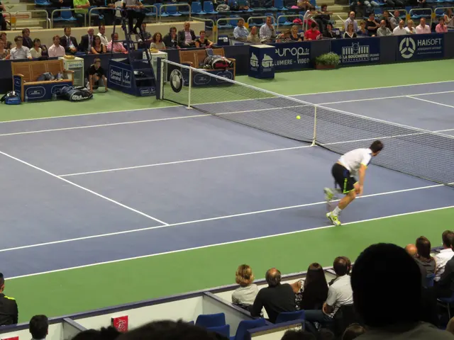 The image shows a man playing tennis on a court in front of a crowd of people sitting in chairs. He...