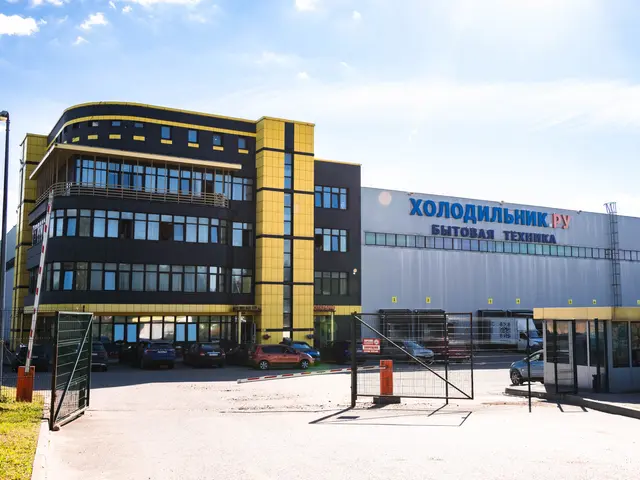 The image shows a large building with a lot of cars parked in front of it, surrounded by a metal...