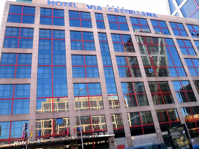 The image shows the Hotel Via Castellana in Madrid, Spain. It is a large building with glass...