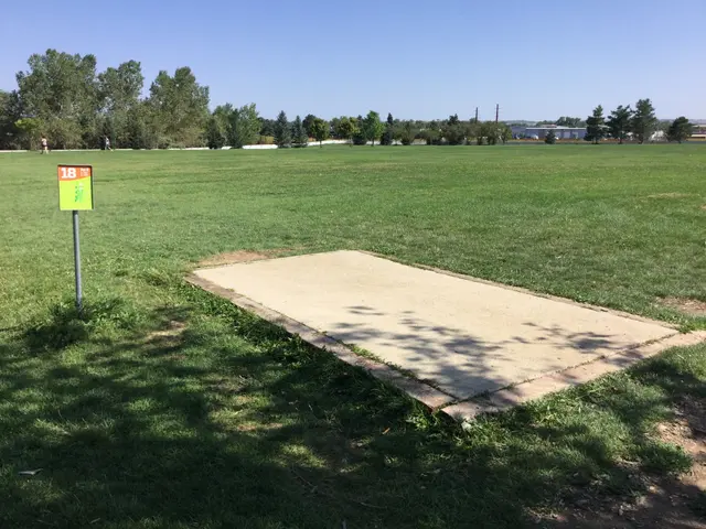 The image shows a grassy field with a sign in the middle of it, surrounded by trees and buildings...