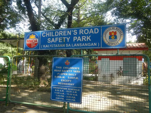 The image shows a children's road safety park with a metal fence surrounding it, two blue boards...