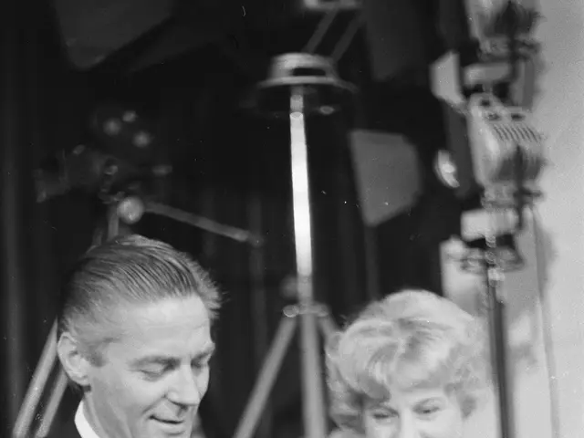 The image shows a man and woman sitting next to each other, both dressed in formal attire. The man...