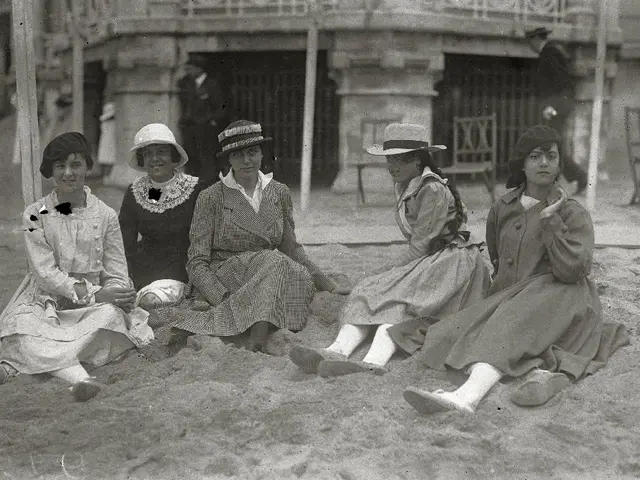 The image shows a group of women sitting on top of a sandy beach, wearing hats and footwear. In the...