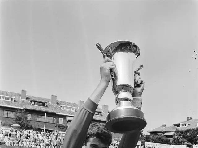 The image shows a man standing on a tennis court, holding up a trophy in triumph. He is surrounded...