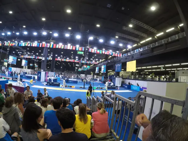 The image shows a large crowd of people sitting in front of a stage, with a railing on the right...