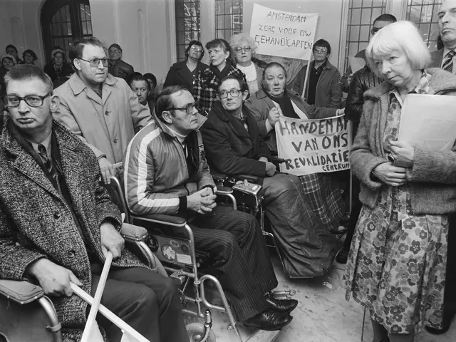 The image shows a group of people sitting in wheelchairs in front of a crowd, some of them holding...