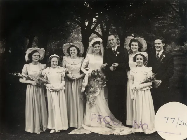 The image shows a black and white photo of a bride and groom surrounded by their wedding party, all...