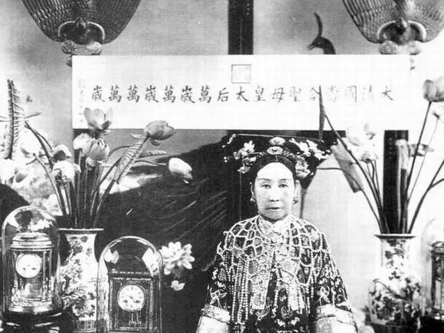 The image shows an old black and white photo of a woman in a kimono standing in front of a table...