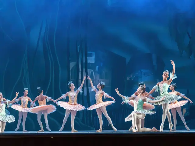 The image shows a group of ballet dancers on a stage in front of a blue background, performing the...