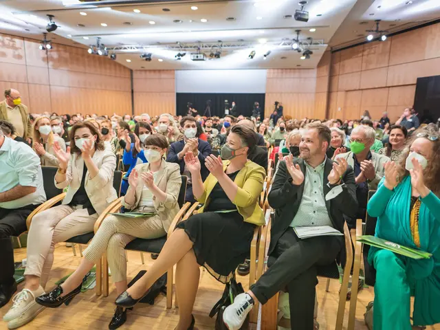 The image shows a group of people sitting in chairs in front of a crowd, clapping their hands in...