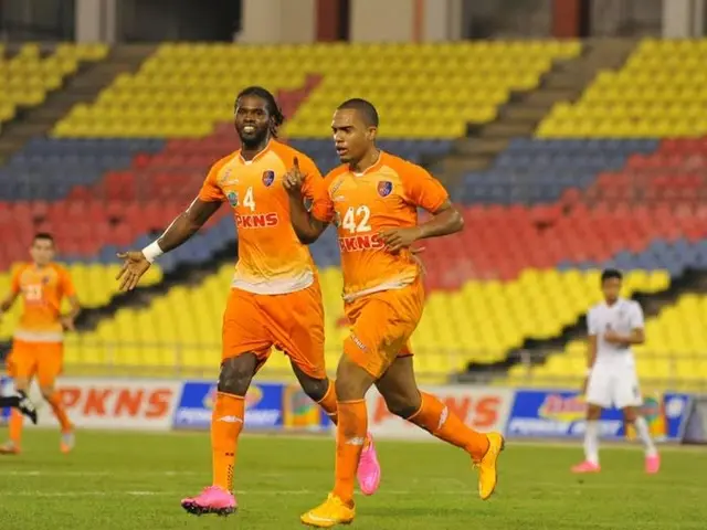 The image shows a group of men playing soccer on a grassy field, with advertisement boards, chairs,...