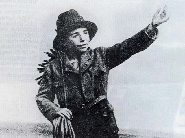 The image shows an old black and white photo of a man in a cowboy outfit, wearing a hat and holding...