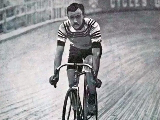 The image shows an old black and white photo of a man riding a bicycle on a track, with a fence and...